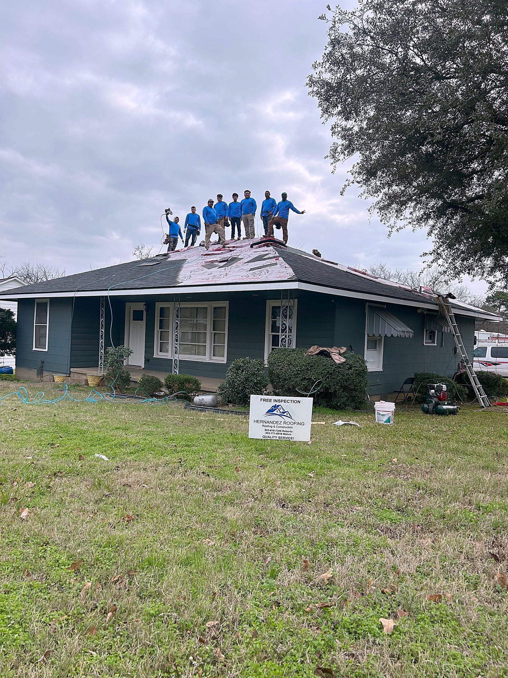 A crew in blue shirts standing along the ridge of a single story house, with a company yard sign staked in the lawn below.