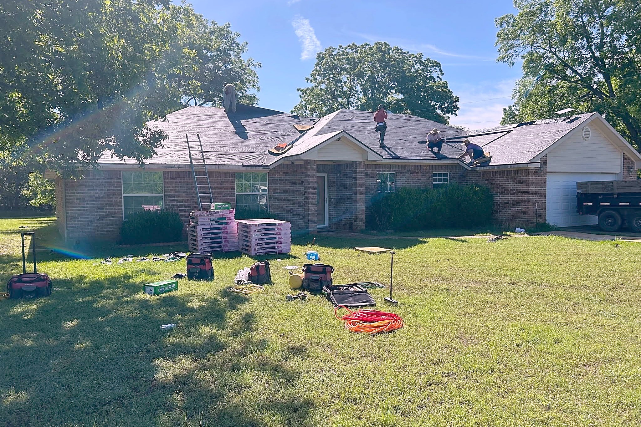 Four roofers working across the roof of a brick ranch house, with bundles of shingles stacked on the lawn and a dump trailer in the drive.