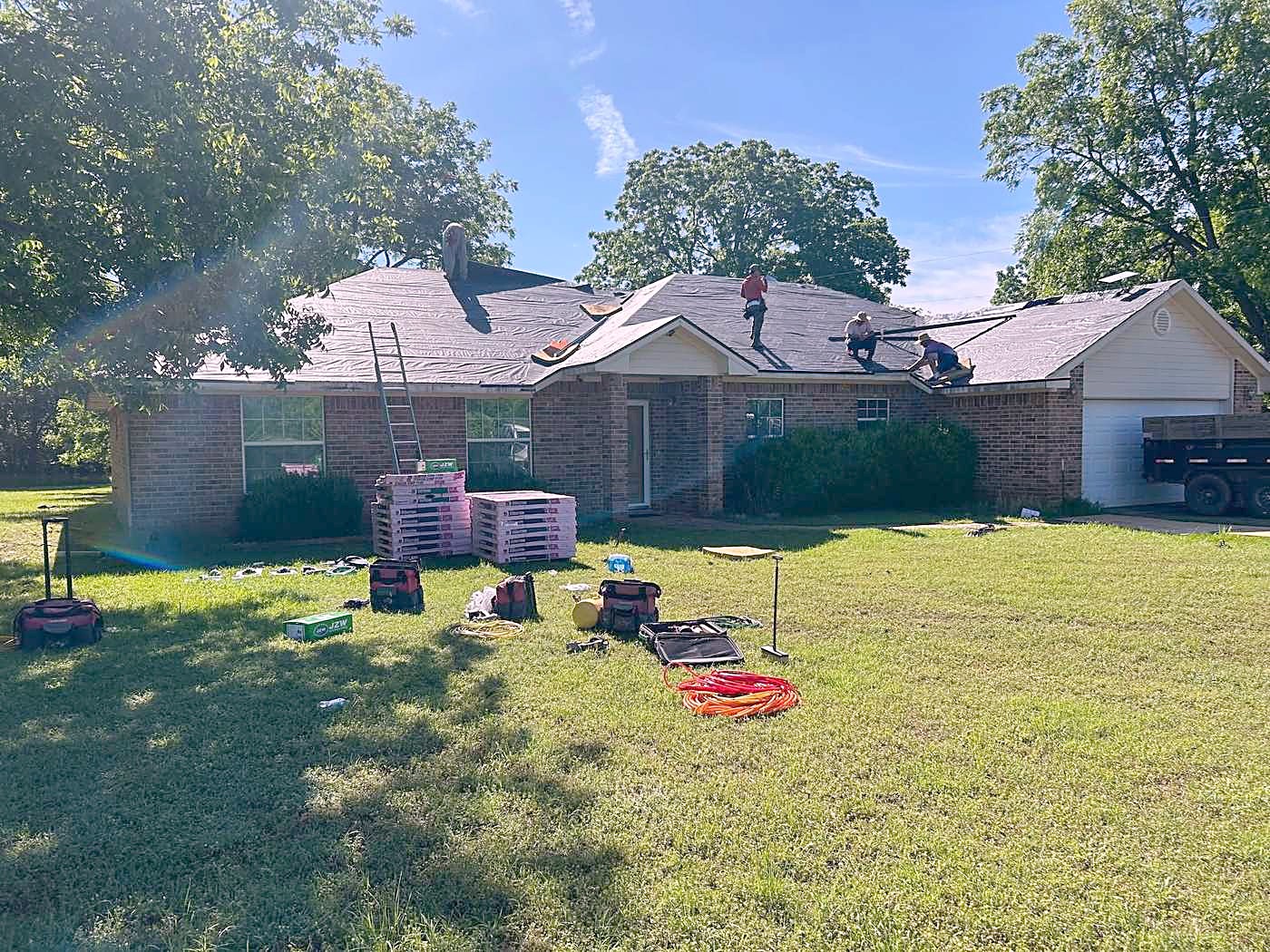 A brick ranch house mid tear off, with a crew on the roof and materials stacked on the lawn.