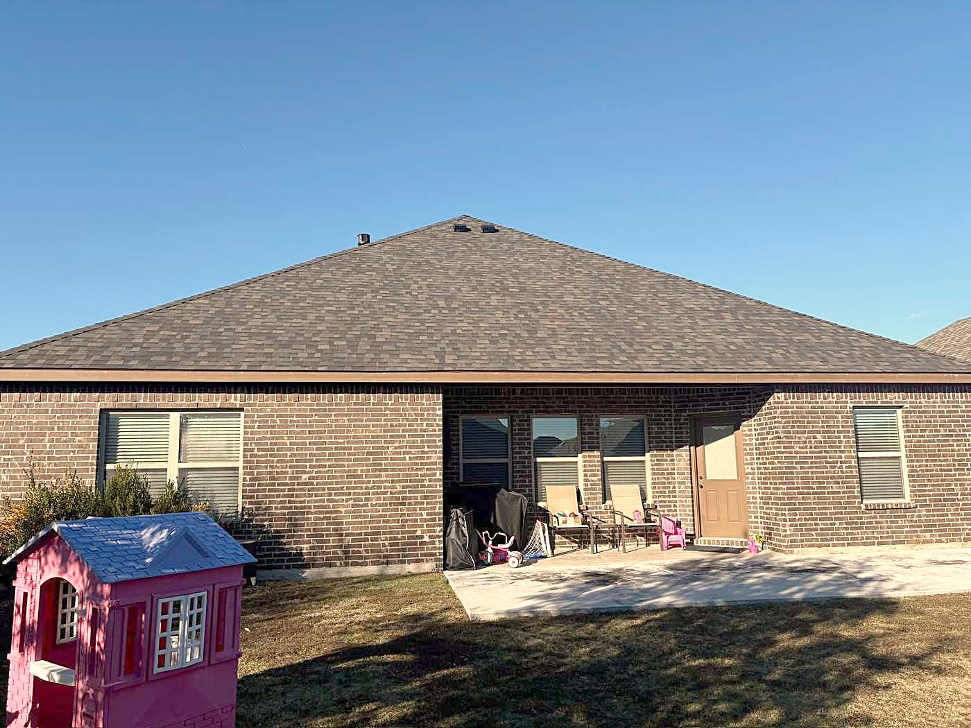 A brick house with its roof finished, seen across the back yard under a blue sky.