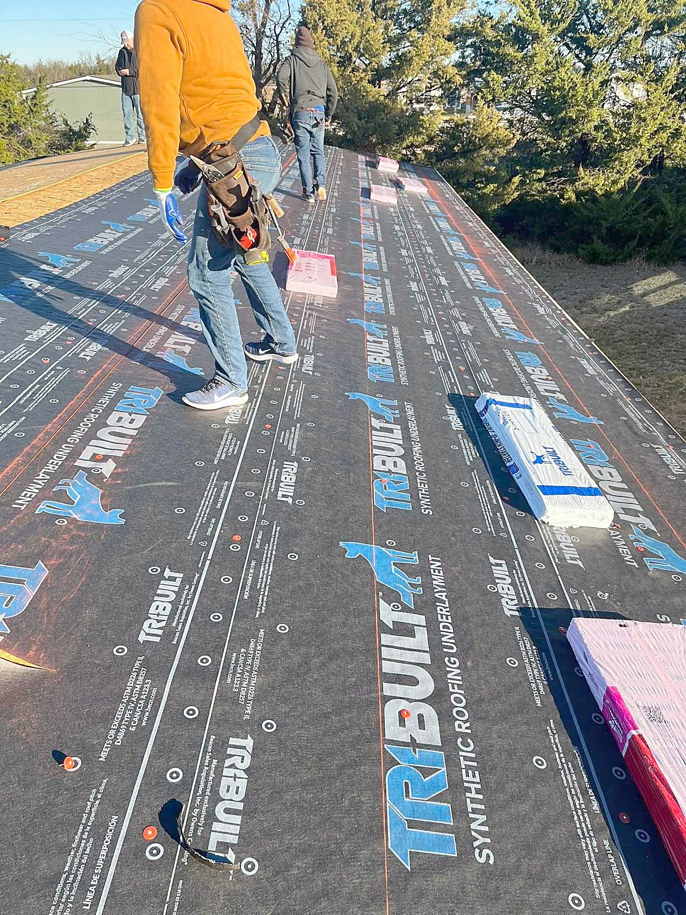 A worker rolling synthetic underlayment across a bare roof deck.